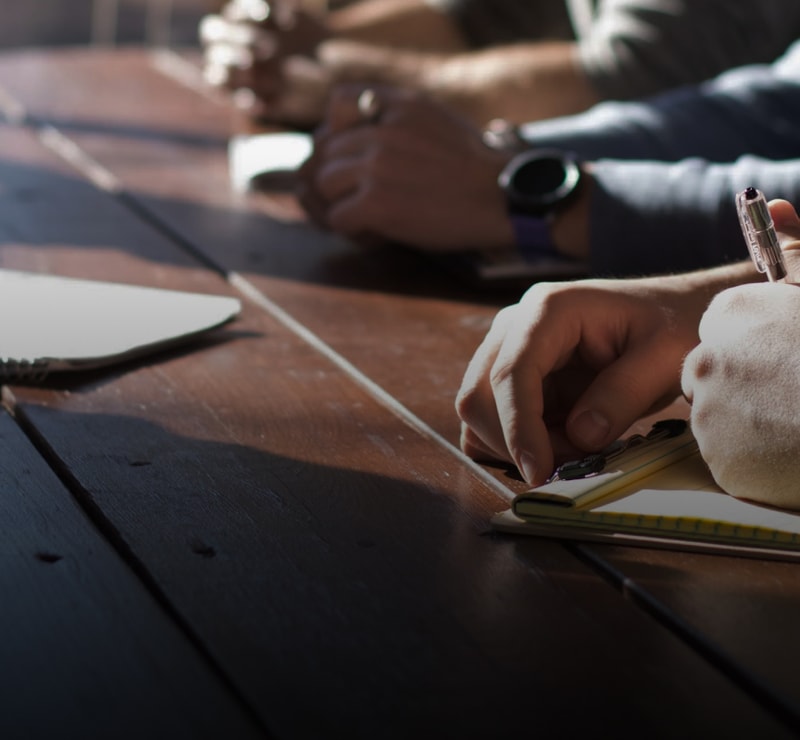 Hands writing with a pen on a notepad at a wooden table, others in the background, a laptop partially visible on the left, warm natural lighting.