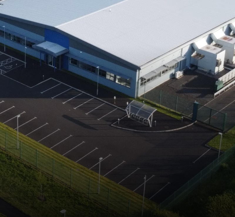 Aerial view of a large industrial building with a light metal roof, adjacent parking lot with white lines, a small curved shelter in the lot, and surrounding fencing and grassy slopes.