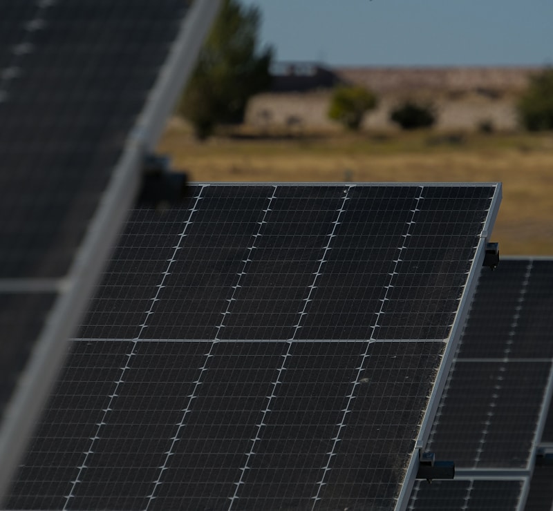 Solar panels in a field, close-up angle showing dark photovoltaic cells with metal framing, distant desert landscape and clear sky in the background.