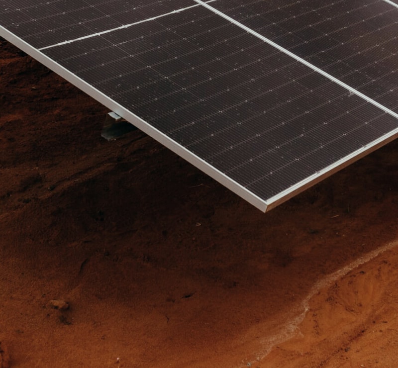Solar panel panel tilted over reddish soil, close-up shot showing dark photovoltaic cells and metallic frame edge intersecting the ground.