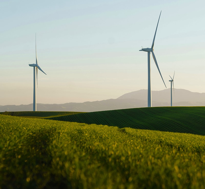 Wind turbines stand tall over rolling green fields with distant hills and a soft sunset glow in the sky.