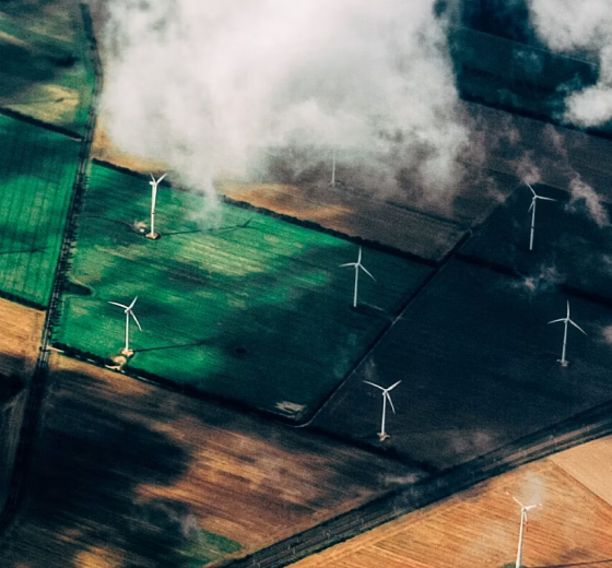 Aerial view of agricultural fields with wind turbines scattered across green and brown plots, fog or low clouds drifting overhead, creating hazy, atmospheric light.