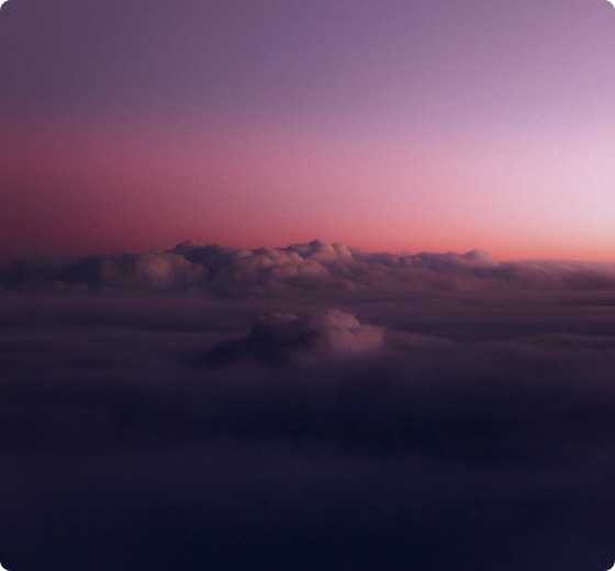 Aerial view of a colorful sunset above a sea of clouds, with soft pink and purple hues along the horizon and darker shadowed cloud layers beneath.