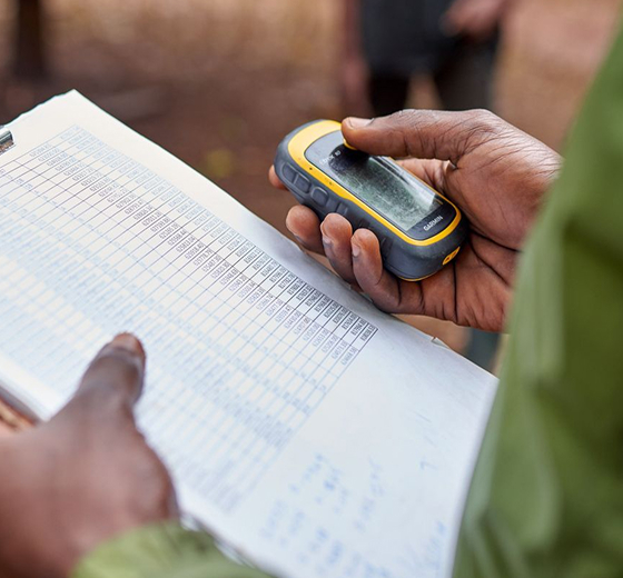 Close-up of a person holding a handheld GPS device over a printed sheet with a dense table, while another hand points at the paper.