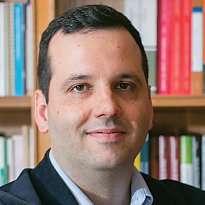 Portrait of a man with short dark hair, wearing a light blue collared shirt and dark blazer, standing in front of a bookshelf with colorful books.