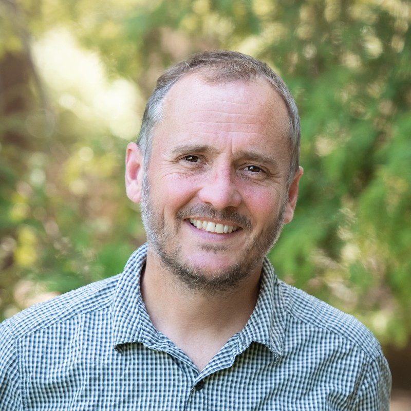 Smiling man with short gray hair and stubble wearing a checkered shirt, outdoors with a blurred green leafy background and natural sunlight.