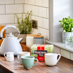 A cozy kitchen scene with a light blue kettle, various mugs on a wooden counter, a box of Yorkshire Tea, and a green plant on the windowsill near subway tile backsplash.