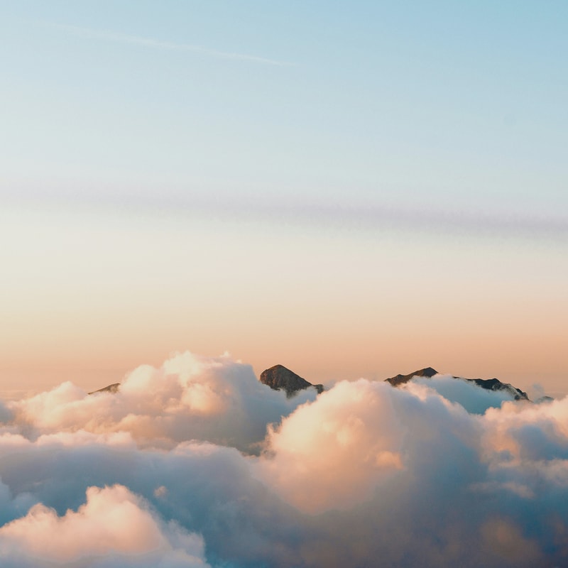 Soft pastel sky above a distant mountain peak peeking through fluffy clouds at sunrise or sunset, with warm peach and cool blue tones.