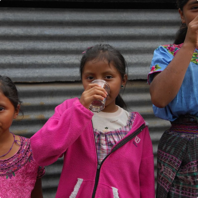 Three smiling children stand against a corrugated metal wall; the central girl in a bright pink fleece drinks from a clear cup, while others wear colorful traditional clothing.