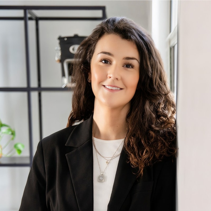 A smiling woman with long, wavy dark hair wearing a black blazer and white top, standing near a window in a modern office space.