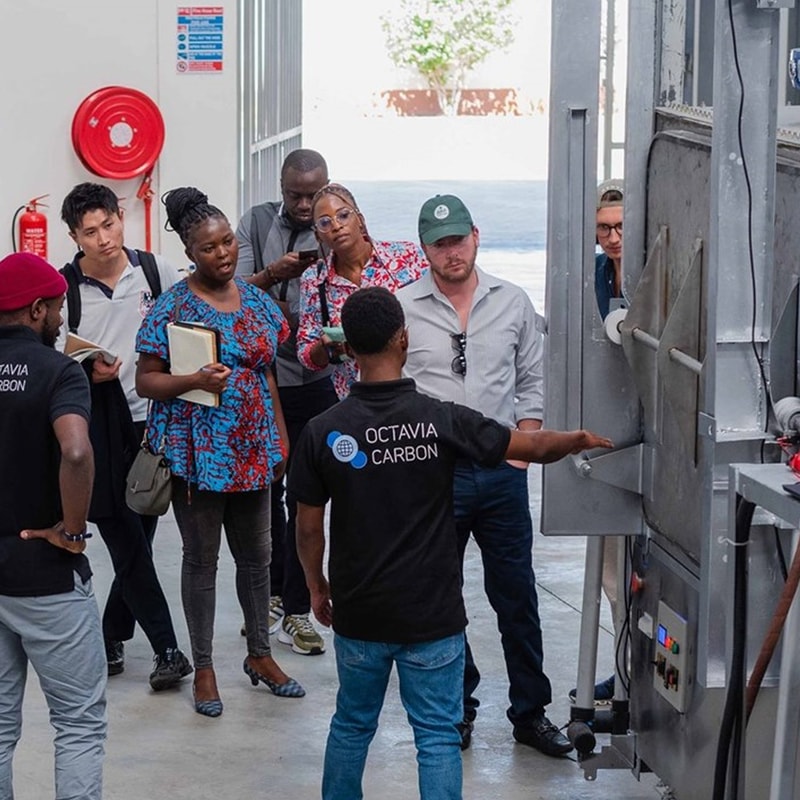 Group of diverse people in a tour or demonstration wearing "OCTAVIA CARBON" shirts, observing a large industrial machine indoors.