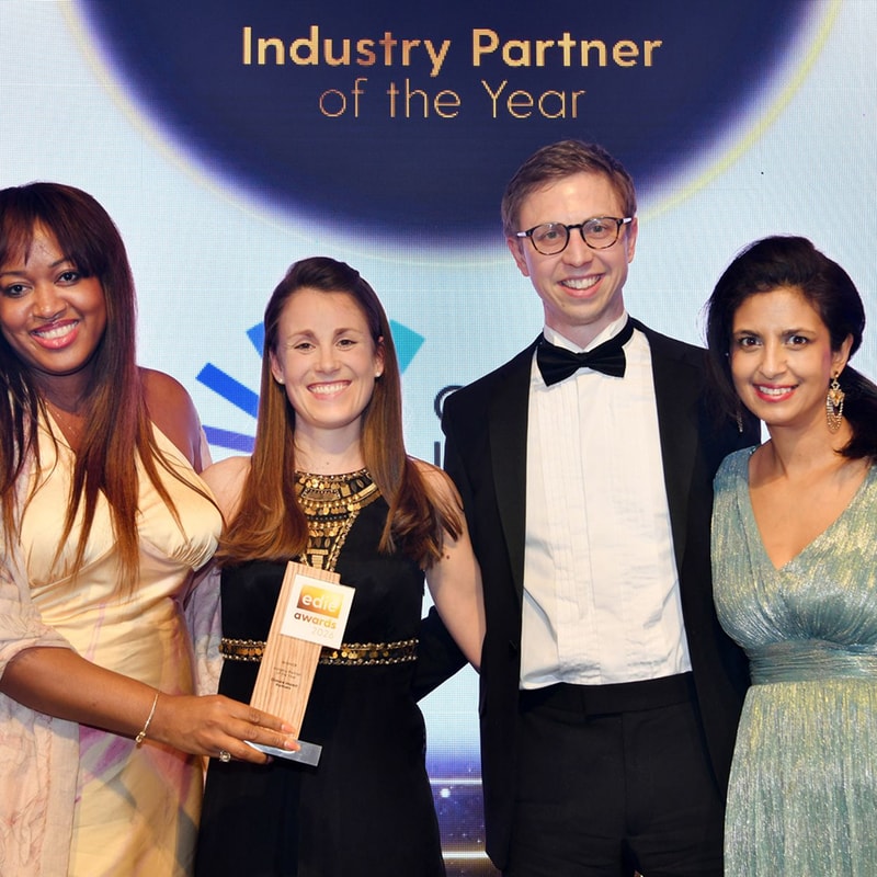 Four people in formal attire pose on stage; a woman in a black dress holds an award, flanked by two women and a man in a tuxedo, all smiling.