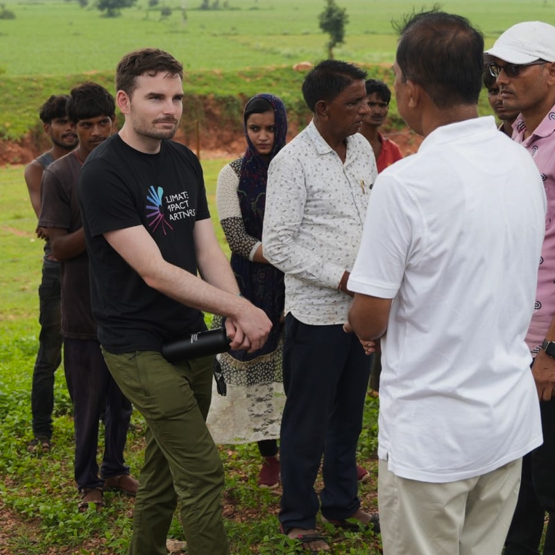 A man in a black T-shirt and olive pants shakes a water bottle while standing in a line of people outdoors, with a green field in the background.