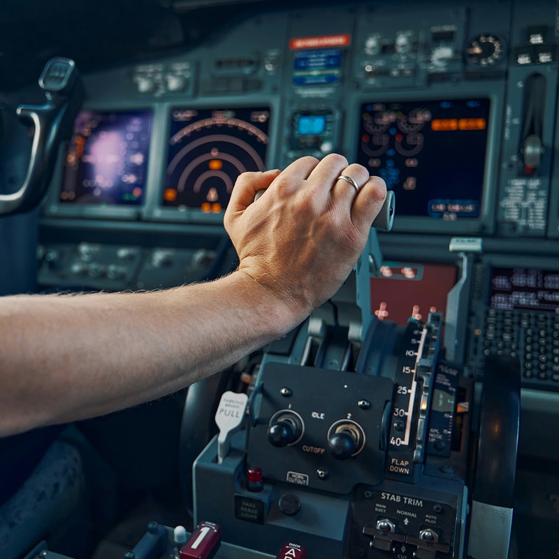Close-up of a hand with a wedding band gripping a Boeing aircraft throttle/control yoke in a cockpit, with digital flight panels and instruments in the blurred background.