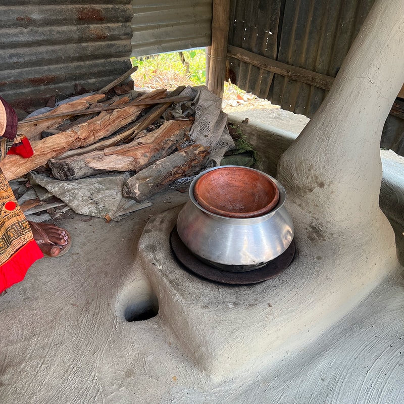 Clay pot sits inside a metal outer container on a clay fireplace stove with a chimney-like structure; a pile of firewood and a corrugated wall in the background.