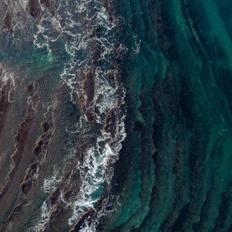 Aerial view of rocky shoreline with turquoise to deep blue waves crashing along stepped rock formations, white foam outlining the coast.