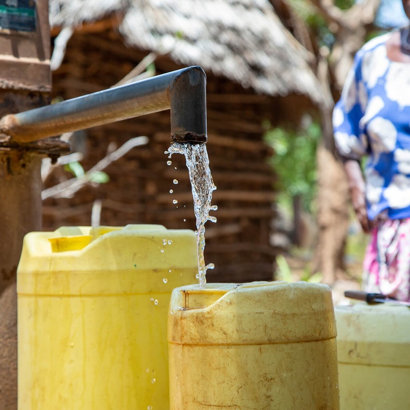 Water pours from a rusted spout into a yellow plastic container, with a muddy outdoor setting and a person in a blue-and-white outfit blurred in the background.