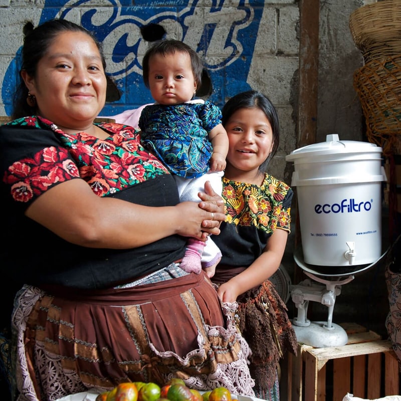 Three people of Central American descent stand together indoors: a smiling woman holding a baby, with a young girl beside them, a water dispenser nearby.