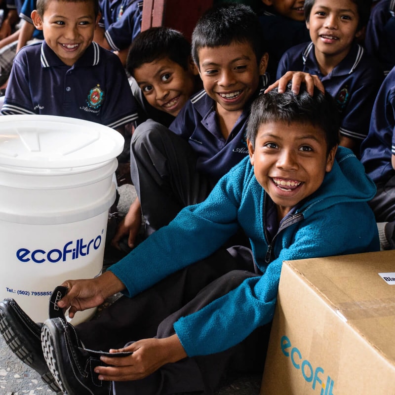Group of smiling children in navy uniforms gathered around a large white EcoFiltro bucket and a cardboard box, one boy in a teal jacket sits at the front.