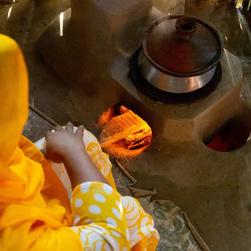 A person in a yellow garment is tending a wood fire in a metal stove, with a glowing orange flame visible through a small opening and a large round stovetop burner nearby.