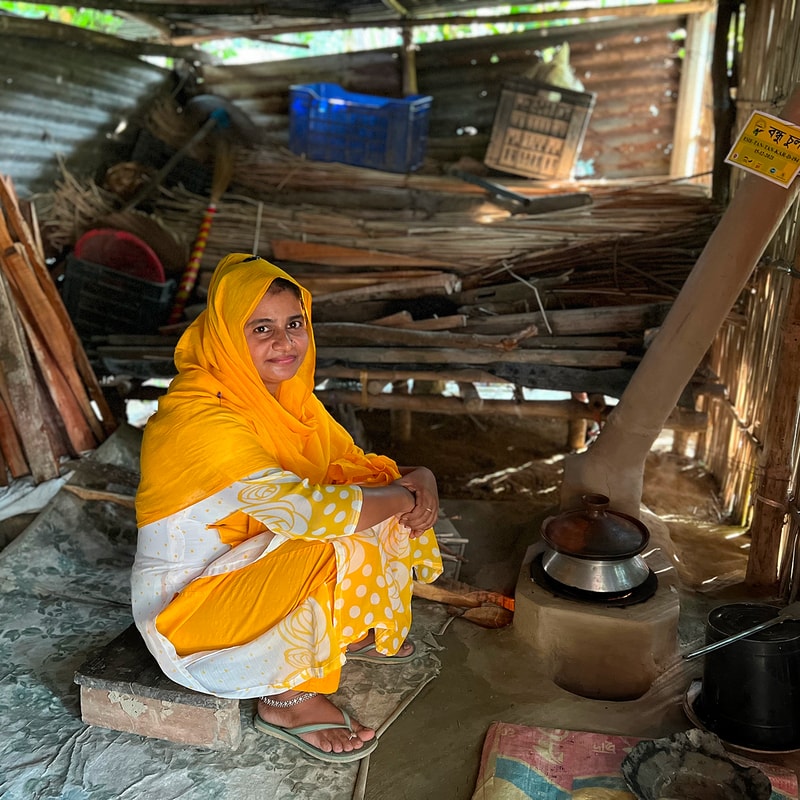 A woman in a bright yellow headscarf and matching outfit sits on a low platform inside a rustic, cluttered kitchen with a stove and pots nearby.