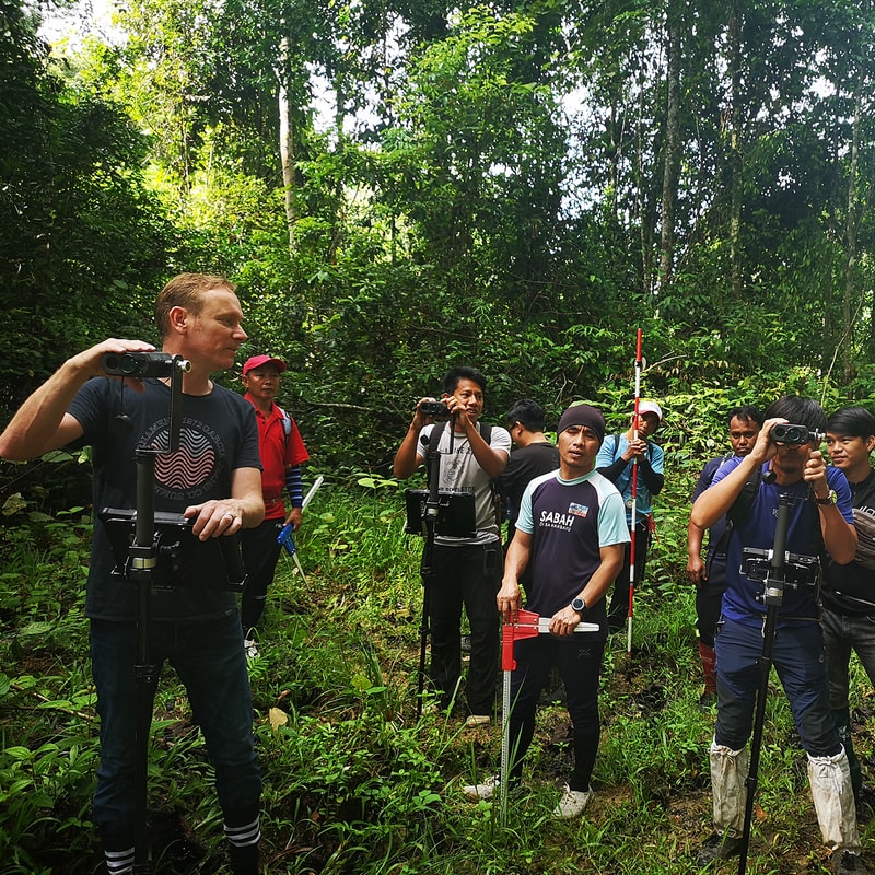 Group of people in a dense forest, some holding binoculars or surveying equipment, one person with a red tool, all focused on the scene ahead.