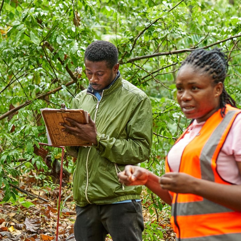 Two people in a lush green outdoor setting: a man in a green jacket writes on a clipboard, while a woman in a bright orange safety vest speaks nearby.