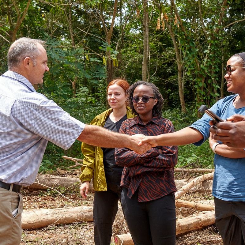A group of four people outdoors in a wooded area, shaking hands between a light-skinned man in a button-down shirt and a person wearing a blue shirt, with two others watching.