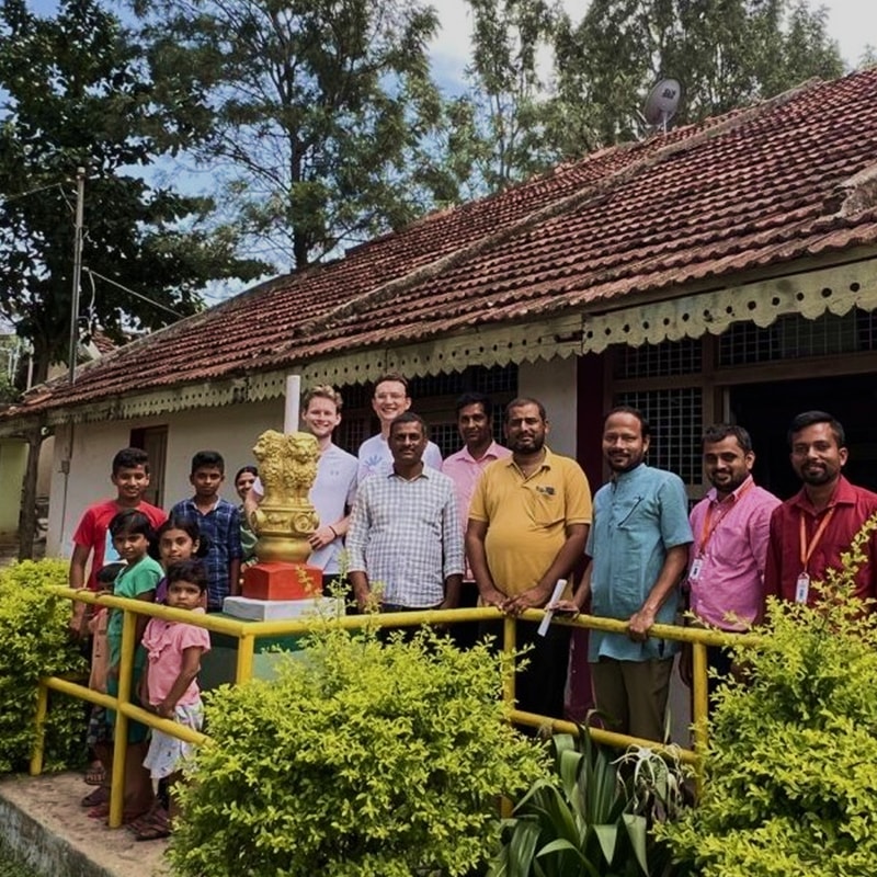 Group of people, including adults and children, posing outdoors in front of a tiled roof building with a decorative statue on a pedestal and yellow railing.