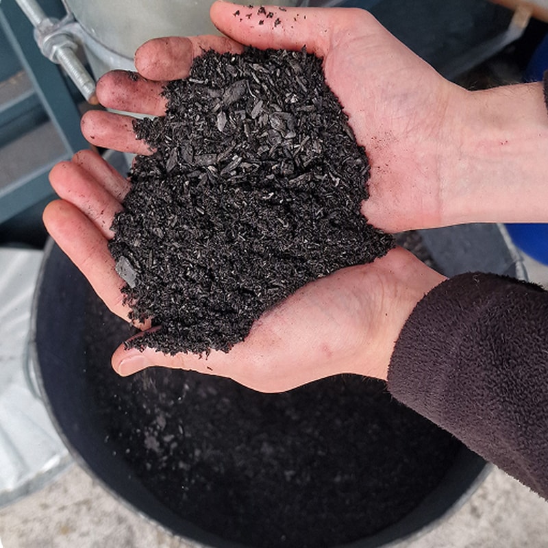 Hands cupping a handful of black shredded material, small glossy pellets and dust, over a dark container; background shows part of industrial equipment.