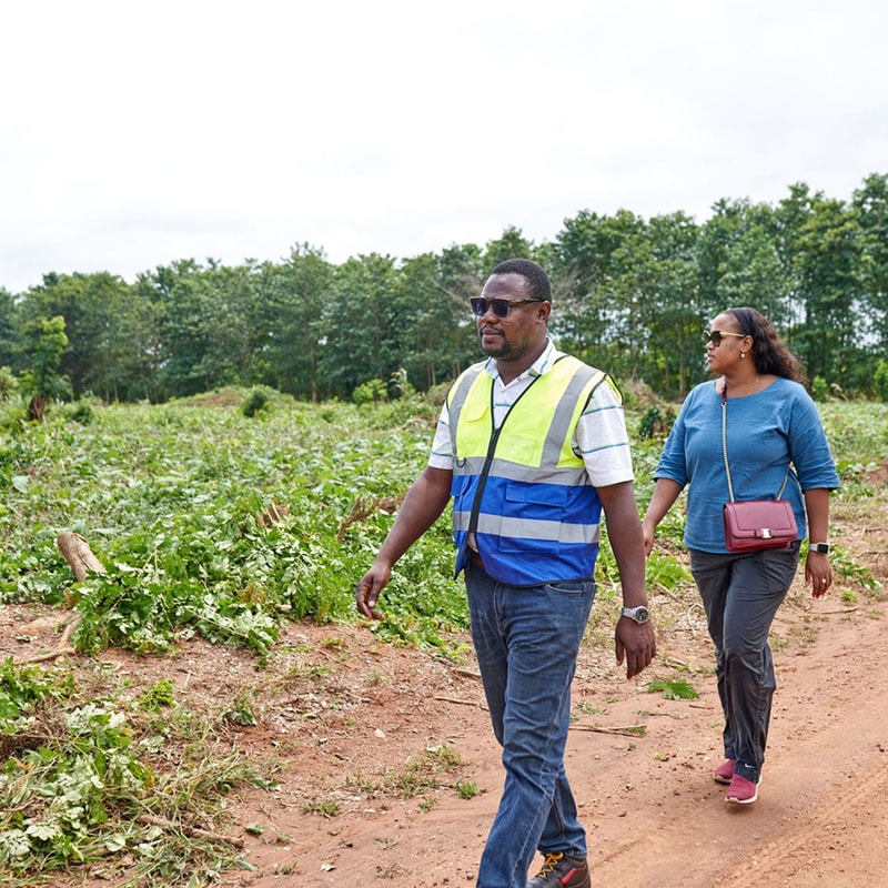 Two people walk along a dirt path in a field; a man in a neon-yellow reflective vest and sunglasses leads, a woman in a blue top and crossbody bag follows.