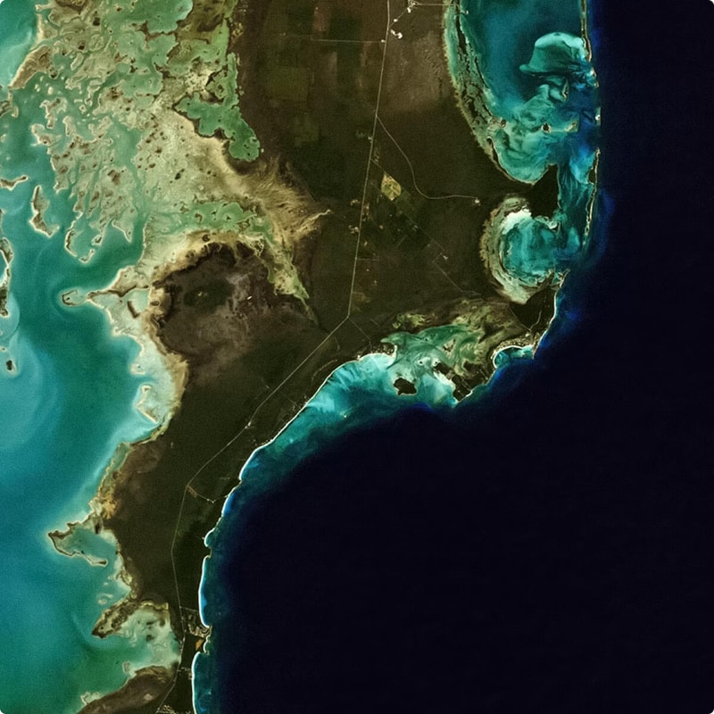 Satellite view of a coastal region with turquoise and emerald shallow waters, dark landmass, and winding roads along a narrow peninsula and sea cliffs.