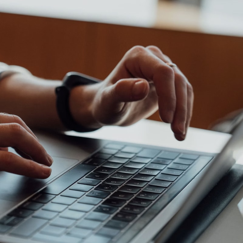 Person typing on a laptop keyboard, with one hand hovering over the keys and wearing a black wristwatch. Close-up urban workspace lighting in background.