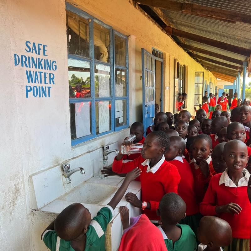 Children in red uniforms line up at a school water point with taps and a labeled sign reading “SAFE DRINKING WATER POINT” along a sunny yellow building.