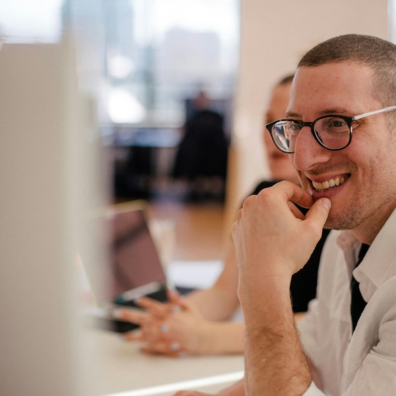 A smiling person with short hair and glasses rests their chin on their hand while looking to the side in a bright office. Another person in the background uses a laptop.