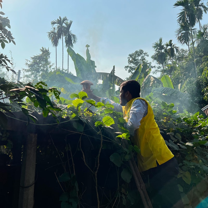 Two people in a lush garden clearing smoke; one wearing a bright yellow vest leans on a wooden fence among dense green vines and tropical trees under a sunny sky.