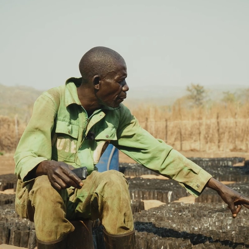 A man wearing a light green work shirt and khaki pants sits among rows of black crates outdoors, pointing to something off to the right while holding a small tool.