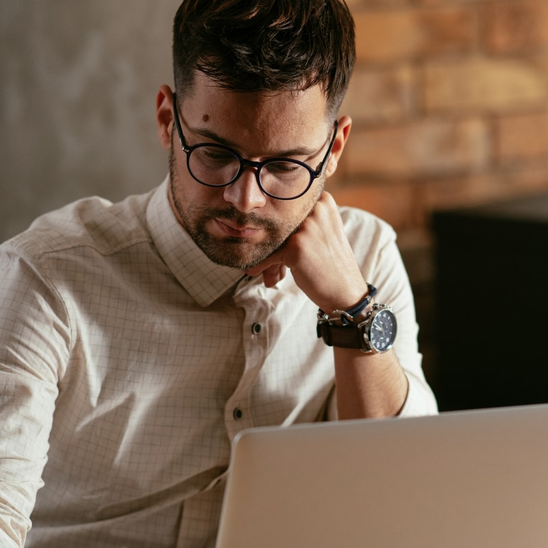 A man with dark hair and glasses sits at a laptop, resting his chin on his hand, wearing a light checkered shirt and a dark wristwatch in a warm, brick-walled setting.