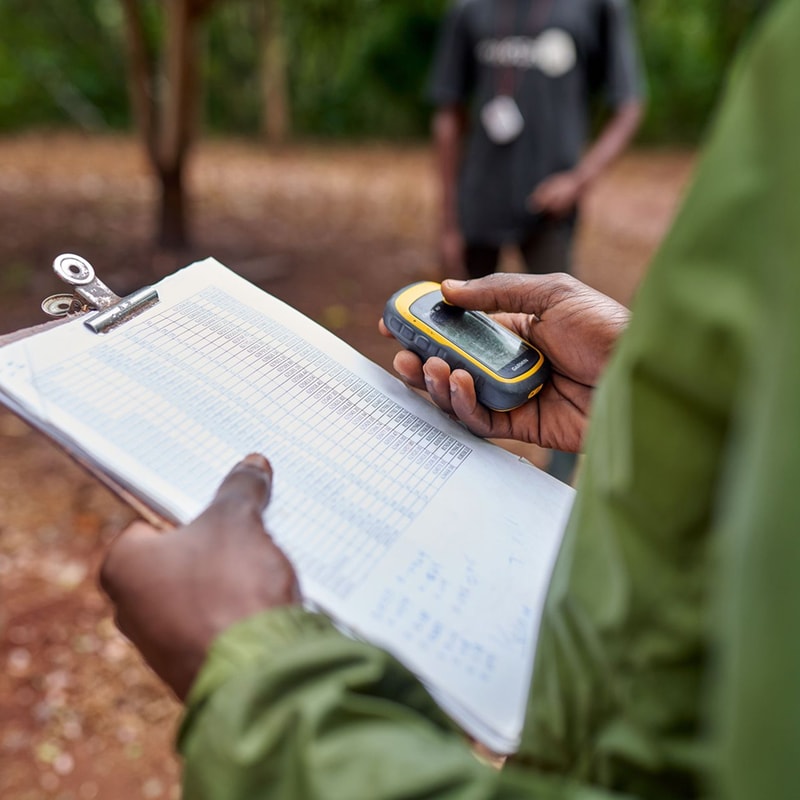 Person holds a clipboard with a chart and uses a handheld GPS device; another person stands in the blurred background outdoors.