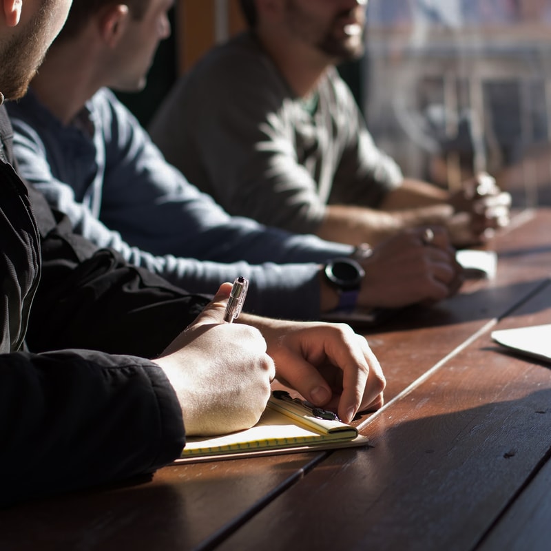 Several people sit at a wooden table, one in the foreground writes in a small notebook with a pen while others blur in the background, pens and hands visible.