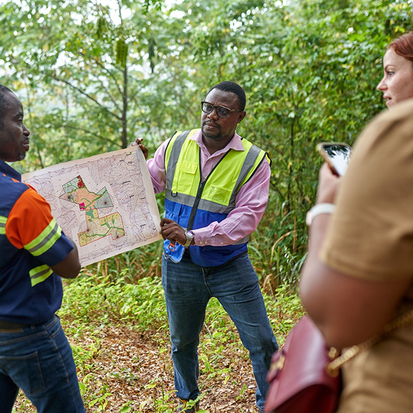 Three adults in high-visibility vests stand in a wooded area, examining a large map together; one points at the map while two onlookers observe, with greenery in the background.