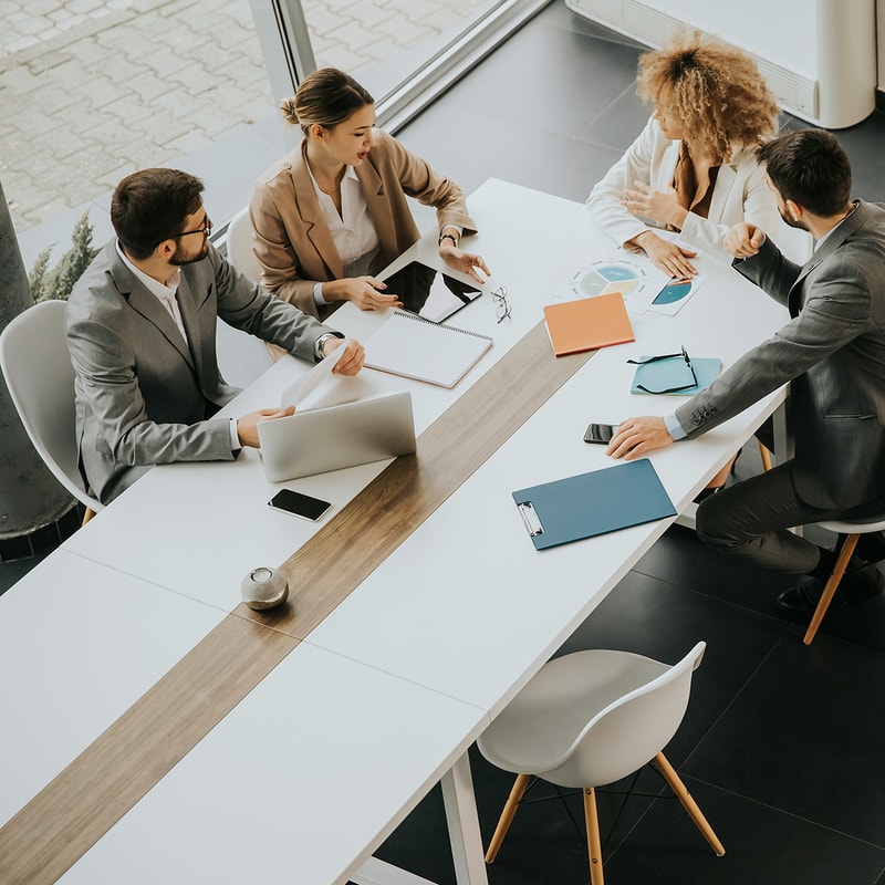 Four professionals in business attire sit around a long white conference table with laptops, notebooks, charts, and smartphones, collaborating in a modern office.