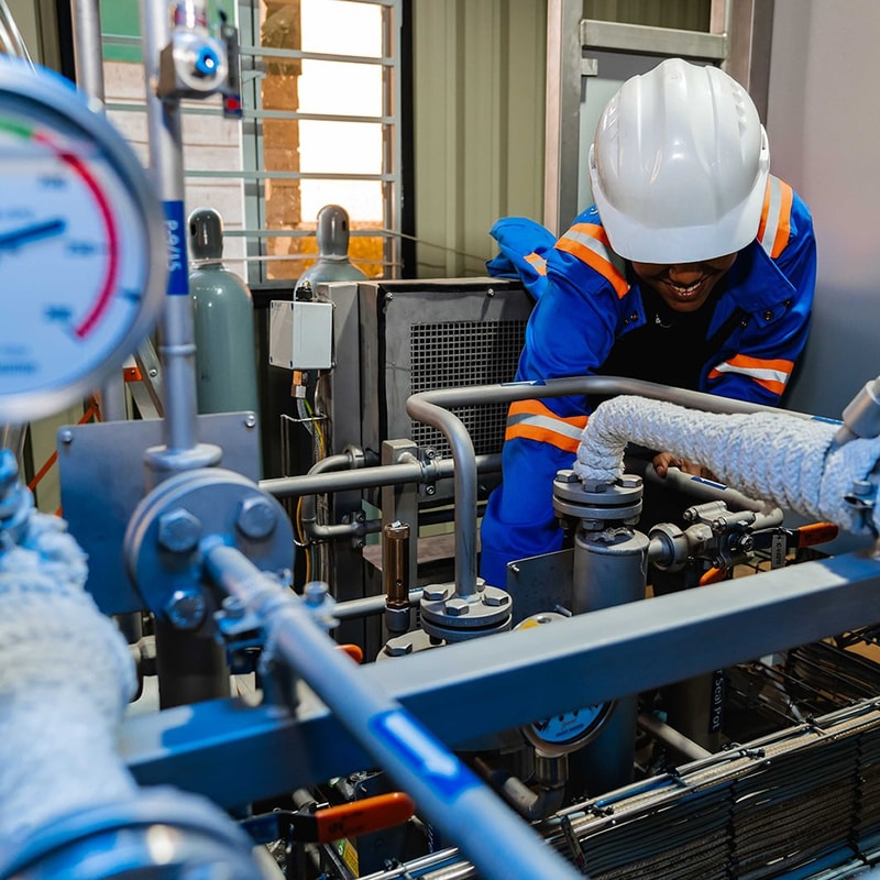 A worker in a white hard hat and blue safety suit with orange stripes leans over industrial pipes and valves inside a mechanical facility.