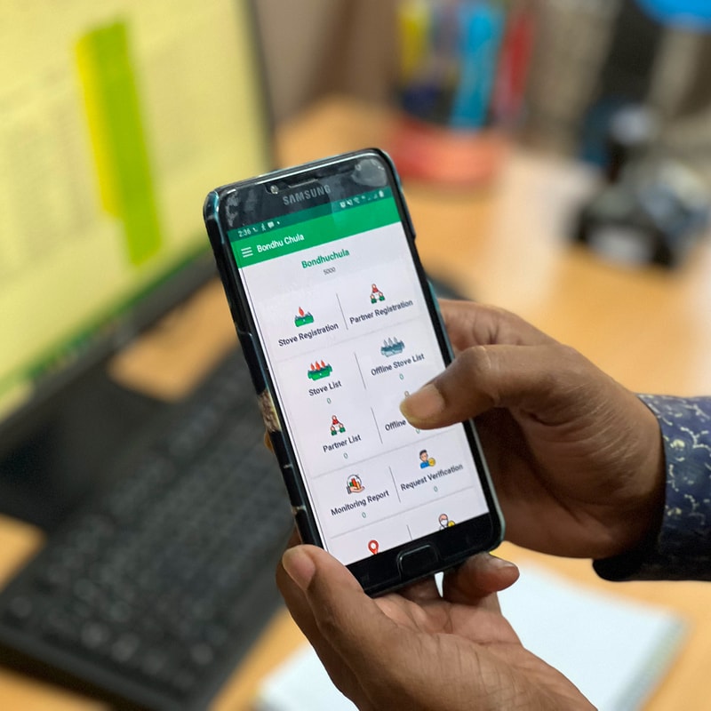 Person holding a Samsung smartphone displaying a green-themed app with menu options such as Stove Registration, Partner Registration, and Online Stove List on a desk near a keyboard.