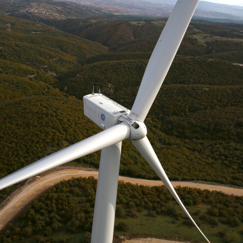 Aerial view of a large white wind turbine with three blades over a green, hilly landscape; a winding dirt road runs beneath, with distant hills in the background.