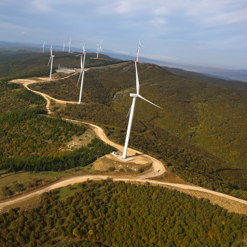 Aerial view of a wind farm on rolling hills with tall white wind turbines along dirt roads, forested terrain, and a distant valley under a cloudy sky.