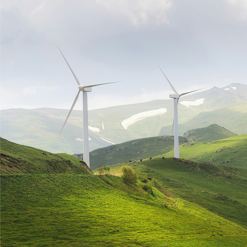 Two tall white wind turbines on green rolling hills with distant mountains, scattered patches of snow, and a cloudy sky in the background.