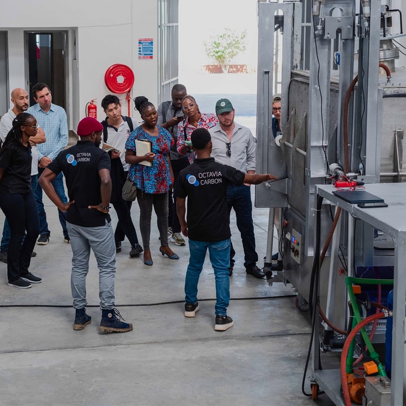 Group of diverse people observing a large industrial machine in a bright lab or facility, some wearing Octavia Carbon shirts, with a presenter explaining details.