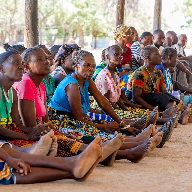A group of people sit side by side outdoors on the ground, many with colorful patterned clothing, barefoot, under a shaded shelter, focusing on something ahead.
