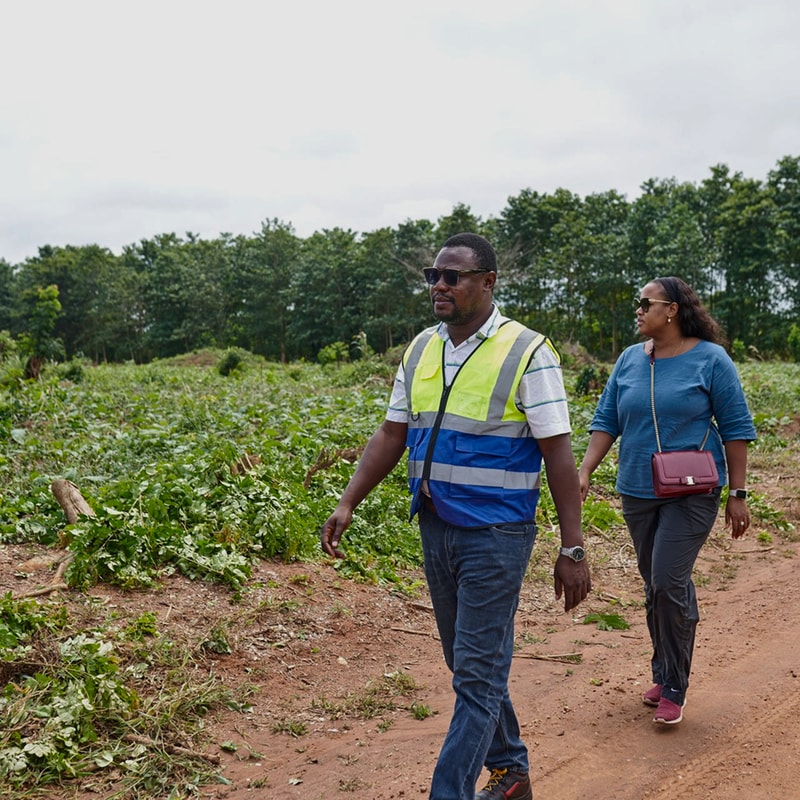 Two people walk along a dirt path in a field of green plants. The man in a high-visibility vest and sunglasses leads, while the woman follows with a burgundy crossbody bag and sunglasses.